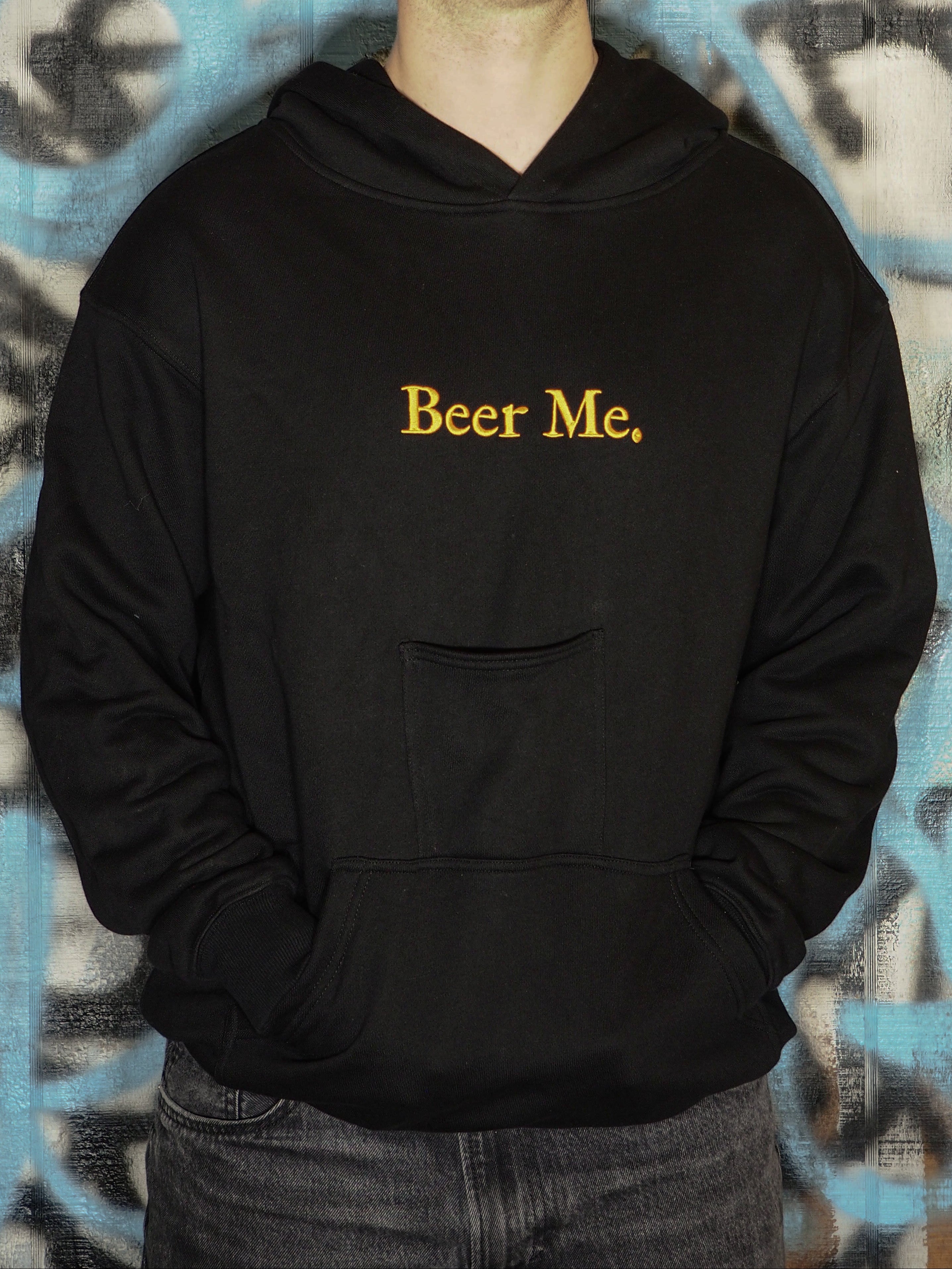 Person wearing a black hoodie with 'Beer Me.' text against a graffiti-covered wall.