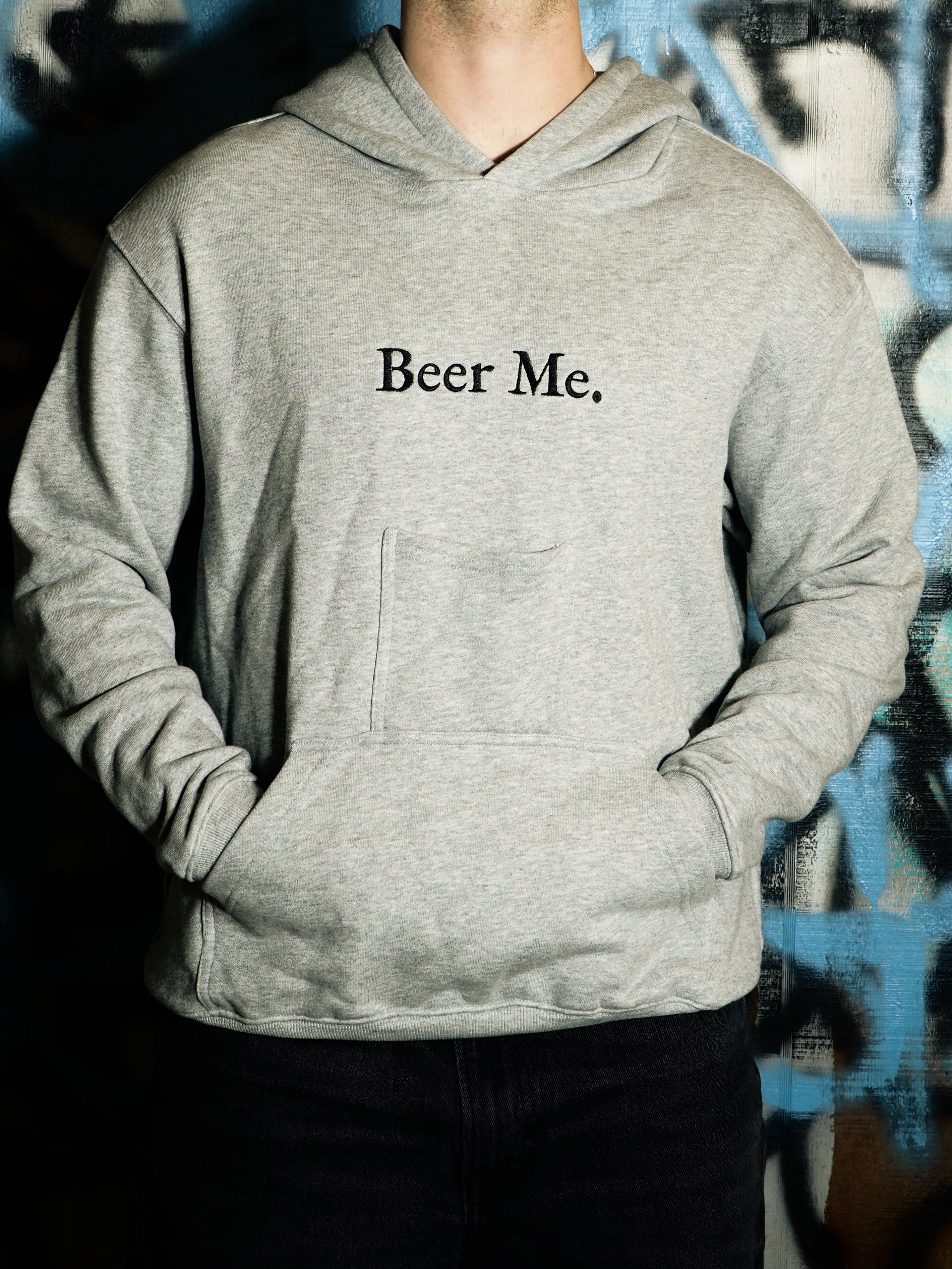 Person wearing a gray hoodie with 'Beer Me.' text against a graffiti-covered wall.